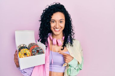 Young hispanic woman with curly hair holding tasty colorful doughnuts box smiling happy pointing with hand and finger 