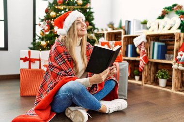 Genç sarışın kadın kitap okuyor. Yılbaşı ağacının yanında oturuyor.