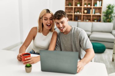 Genç beyaz çift dizüstü bilgisayar kullanıyor ve kahve içiyor. Evdeki masada oturuyorlar..