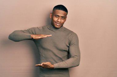 Balıkçı yaka kazak giyen genç siyahi bir adam. Elleriyle büyük ve büyük bir işaret, ölçü sembolü gösteriyor. Kameraya bakıp gülümsüyor. ölçüm kavramı. 