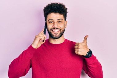 Sakallı genç Arap adam akıllı telefonla konuşuyor mutlu ve olumlu gülümsüyor, başparmağını kaldırıyor ve onaylıyor. 
