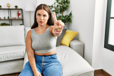 Evdeki kanepede oturan genç esmer kadın parmağını kaldırmış ve kızgın bir ifadeyle işaret ediyor. 