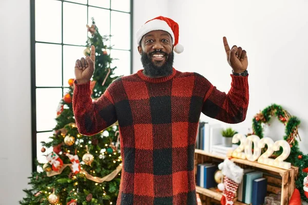 アフリカ系アメリカ人男性身に着けているサンタクラスの帽子立ってクリスマスツリーによって笑顔と驚きと指摘指と腕を上げ — ストック写真