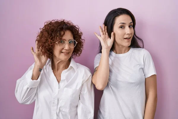 ヒスパニック系の母親と娘ピンクの背景にピンクの背景にカジュアルな白いTシャツを着て手を耳に噂やゴシップに耳を傾け笑みを浮かべて 聴覚障害の概念 — ストック写真