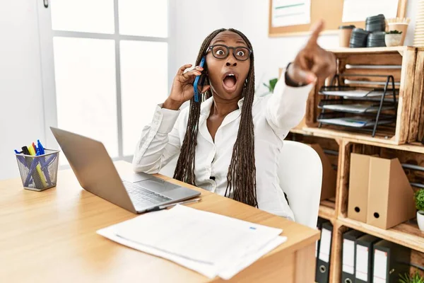 Black woman with braids working at the office speaking on the phone pointing with finger surprised ahead, open mouth amazed expression, something on the front