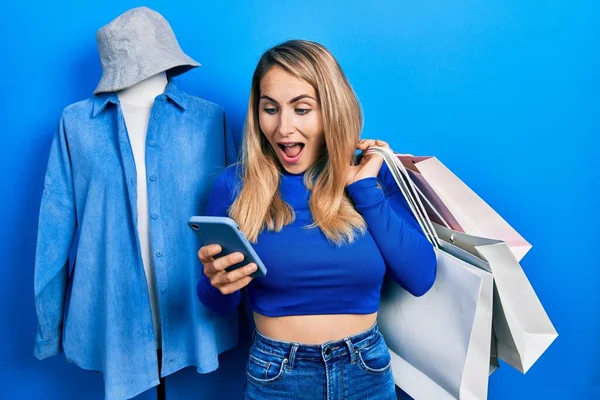 Young caucasian woman holding shopping bags and smartphone celebrating crazy and amazed for success with open eyes screaming excited.