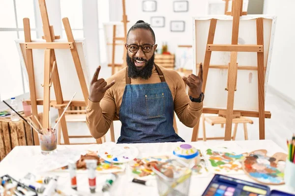 African american artist man at art studio shouting with crazy expression doing rock symbol with hands up. music star. heavy concept.