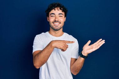 Latin asıllı genç bir adam, gündelik beyaz tişört giyip kameraya hayret ediyor ve gülümsüyor.. 