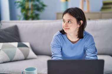 Down sendromlu genç bir kadın dizüstü bilgisayar kullanıyor ve evdeki kanepede oturup kahve içiyor.