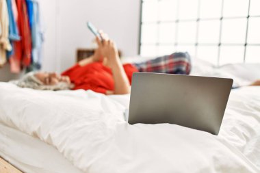 Orta yaşlı, kır saçlı, yatak odasındaki yatakta uzanmış akıllı telefon kullanan bir kadın..
