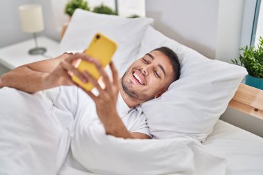 Genç İspanyol adam yatakta akıllı telefon kullanıyor.