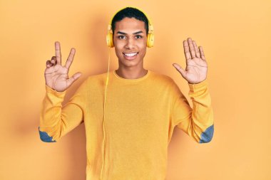 Genç Afro-Amerikalı adam müzik dinlerken kulaklık takıyor ve 8 numaralı parmaklarıyla işaret ederken kendinden emin ve mutlu gülümsüyor.. 