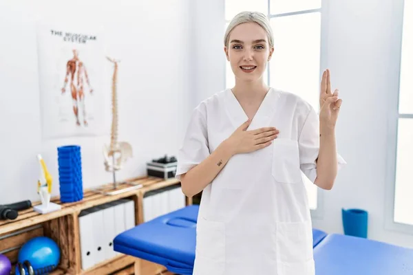 Young caucasian woman working at pain recovery clinic smiling swearing with hand on chest and fingers up, making a loyalty promise oath