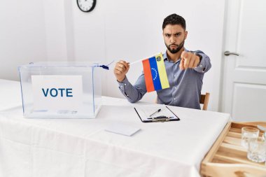 Young handsome man with beard at political campaign election holding venezuela flag pointing with finger to the camera and to you, confident gesture looking serious 