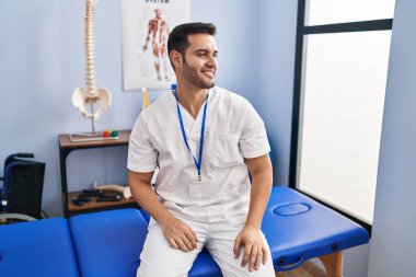 Sakallı, sakallı, acılı bir klinikte çalışan, yüzünde gülümseme ve doğal ifadeyle yan yana bakan genç bir İspanyol. Kendinden emin gülen. 