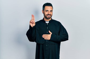 Sarışın, sakallı, Katolik rahip cübbesi giyen, elleri göğsünde, parmakları havada yemin eden, sadakat yemini eden yakışıklı bir İspanyol. 