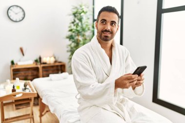 Genç İspanyol bornoz giyen adam güzellik merkezinde akıllı telefon kullanıyor.