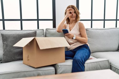Orta yaşlı bir kadın koltukta oturmuş, teslimat paketini kontrol ediyor, gülümsüyor ve gülümsüyor. 