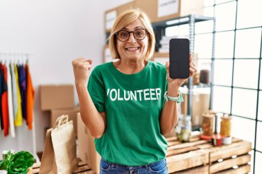 Middle age blonde woman working wearing volunteer t shirt showing smartphone screen screaming proud, celebrating victory and success very excited with raised arms 
