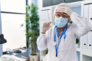 Senior woman with grey hair wearing doctor uniform and medical mask holding syringe stressed and frustrated with hand on head, surprised and angry face 