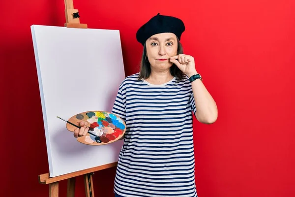Middle age hispanic woman standing drawing with palette by painter easel stand mouth and lips shut as zip with fingers. secret and silent, taboo talking 