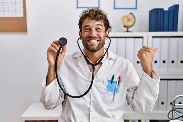 Young hispanic doctor man wearing doctor uniform holding stethoscope at clinic pointing thumb up to the side smiling happy with open mouth