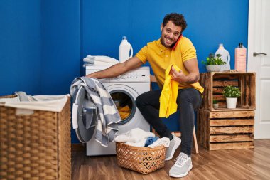 Genç İspanyol adam çamaşır odasında çamaşır yıkayarak akıllı telefondan konuşuyor.