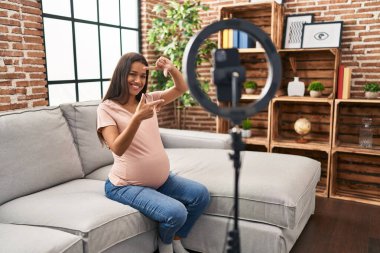 Genç hamile kadın evde akıllı telefonuyla video kaydı yapıyor gülümseyerek elleri ve parmakları gülen yüzüyle çerçeve yapıyor. yaratıcılık ve fotoğrafçılık kavramı. 