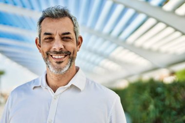 Orta yaşlı, kır saçlı, gülümseyen bir adam şehirde mutlu bir şekilde duruyor..