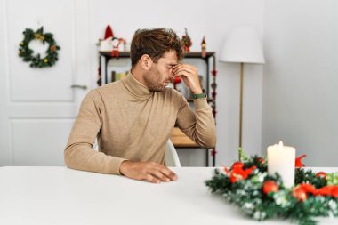 Yılbaşı süslemeleri sırasında masanın üzerinde oturan sakallı yakışıklı genç adam burnuna ve gözlerine sürtünmekten yorgun ve yorgun düştü. Stres ve hüsran kavramı. 