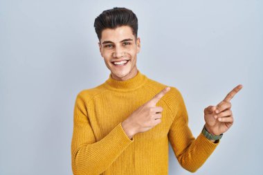 Genç İspanyol adam mavi arka planda duruyor gülümsüyor ve kameraya bakıyor iki eliyle ve parmaklarıyla kenarı işaret ediyor.. 