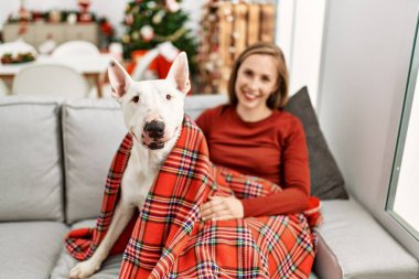 Beyaz bir kadın gülerek evde köpekle oturuyor. Noel ağacı yanında.