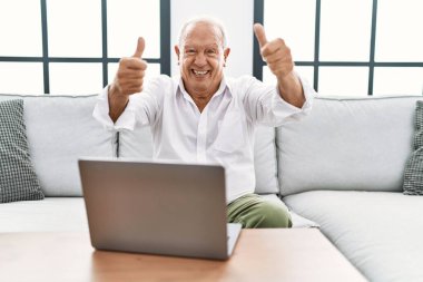 Senior man using laptop at home sitting on the sofa approving doing positive gesture with hand, thumbs up smiling and happy for success. winner gesture. 