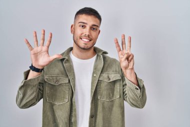 İzole edilmiş arka planda duran genç İspanyol adam 8 numaralı parmaklarıyla gülümseyerek kendinden emin ve mutlu bir şekilde işaret ediyor.. 