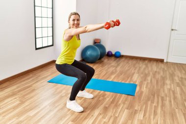 Genç sarışın kız spor merkezinde dambıllar kullanarak mutlu bir antrenman yapıyor..