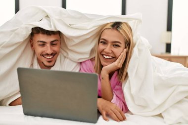 Beyaz tenli genç çift çarşafla örtüyor ve evdeki yatakta dizüstü bilgisayar kullanıyorlar..