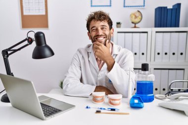 Tıbbi klinikte çalışan genç İspanyol dişçi kameraya kollarını kavuşturup gülümseyerek ve çenesini kaldırarak bakıyor. Olumlu düşünmek. 