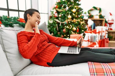 Young hispanic woman having video call sitting by christmas tree at home