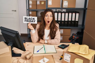 Young brunette woman holding black friday banner small commerce scared and amazed with open mouth for surprise, disbelief face 