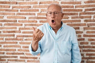 Senior man with grey hair standing over bricks wall surprised pointing with hand finger to the side, open mouth amazed expression. 