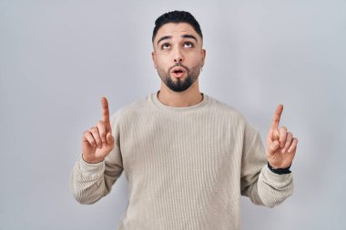 Soyutlanmış arka planda duran yakışıklı genç adam şaşırmış ve şaşırmış bir şekilde yukarı bakıyor ve parmaklarıyla işaret ediyor ve kollarını kaldırıyor.. 