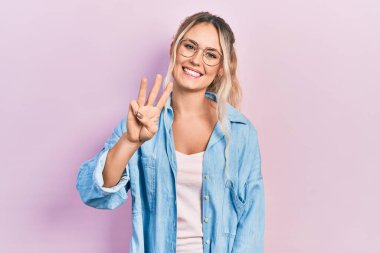 Güzel, genç, sarışın bir kadın. Günlük kıyafetler ve gözlük takıyor. Üç numaralı parmaklarıyla gösteriyor ve işaret ediyor.. 