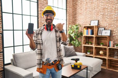 Young hispanic man with beard working at home renovation holding smartphone pointing thumb up to the side smiling happy with open mouth 