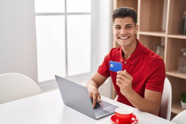 Evdeki masada oturan, dizüstü bilgisayar ve kredi kartı kullanan genç bir İspanyol.