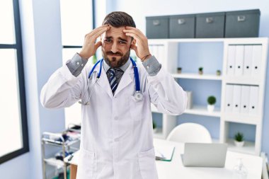 Doktor üniforması giyen yakışıklı İspanyol bir adam ve tıbbi klinikte steteskop. Ağrı ve migren yüzünden çaresiz ve stresli. Eller başının üstünde.. 