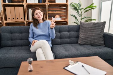 Young brunette woman at consultation office smiling looking to the camera showing fingers doing victory sign. number two. 