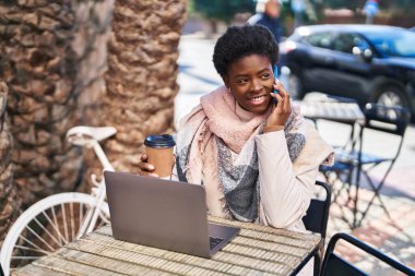 Afro-Amerikalı kadın bilgisayarını kullanıp kahve dükkanının terasında oturmuş akıllı telefondan konuşuyor.