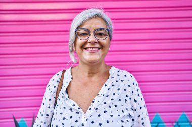 Orta yaşlı bir kadın pembe arka planda kendine güvenerek gülümsüyor.