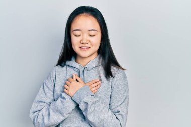 Sıradan kıyafetler giyen genç Çinli bir kız elleri göğsünde, gözleri kapalı ve yüzünde minnet dolu bir jest ile gülümsüyor. sağlık kavramı. 