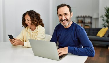 Orta yaşlı İspanyol çift evde bilgisayar ve akıllı telefon kullanarak mutlu bir şekilde gülümsüyor..